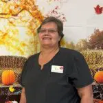 Older woman with gray/black hair and glasses wears dark gray scrubs and smiles for the camera while standing in front of a fall background