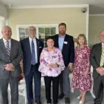 Four men in suits and two women in business attire smile in a row together on a porch in front of rocking chairs and a window