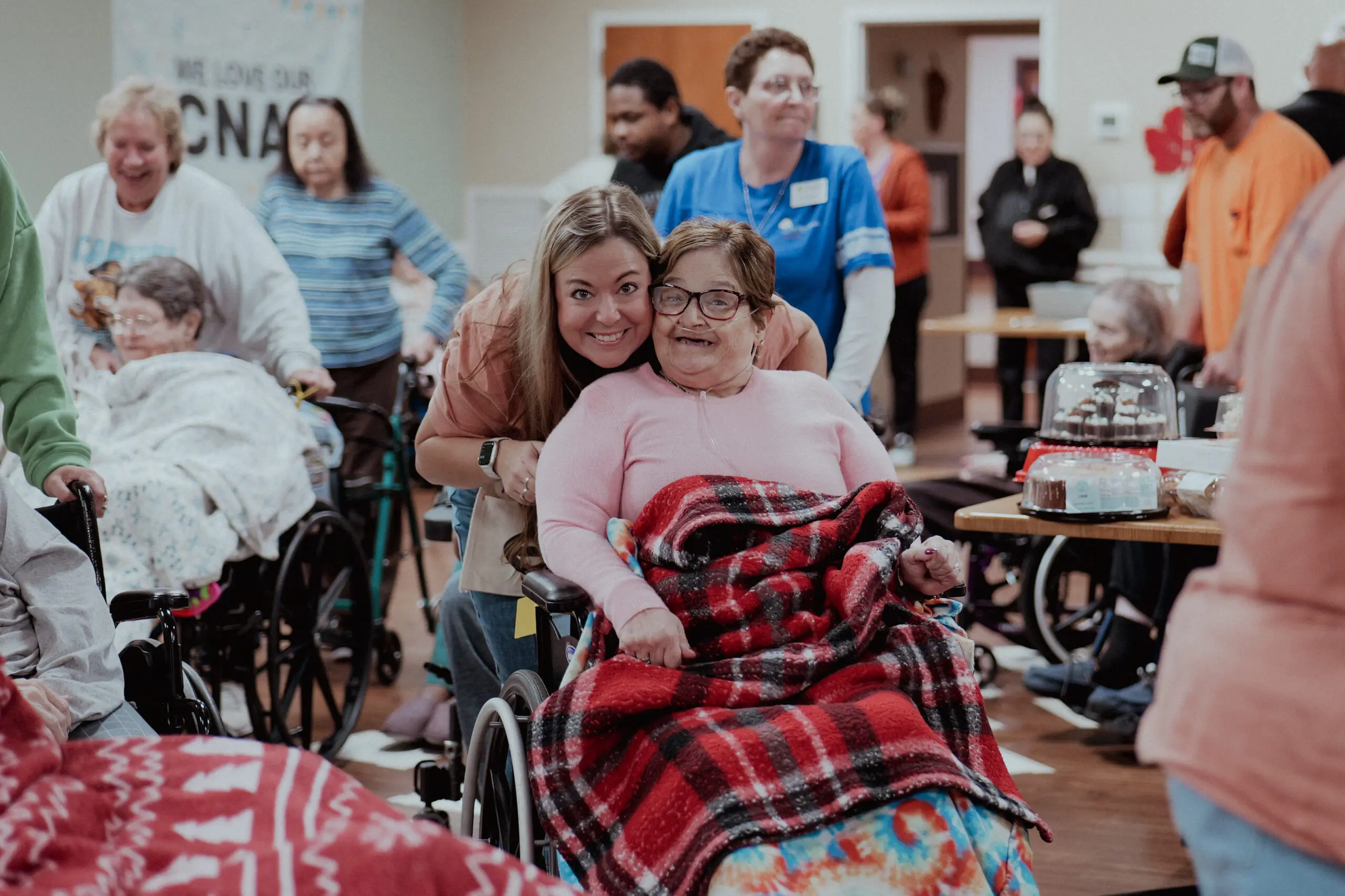 Woman in an orange top smiles with a female patient with an oxygen machine in a wheelchair in a room filled with other patients and caregivers