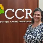 Dark haired woman in a navy and white patterned top smiles in front of the CCR logo in white on a wooden panel