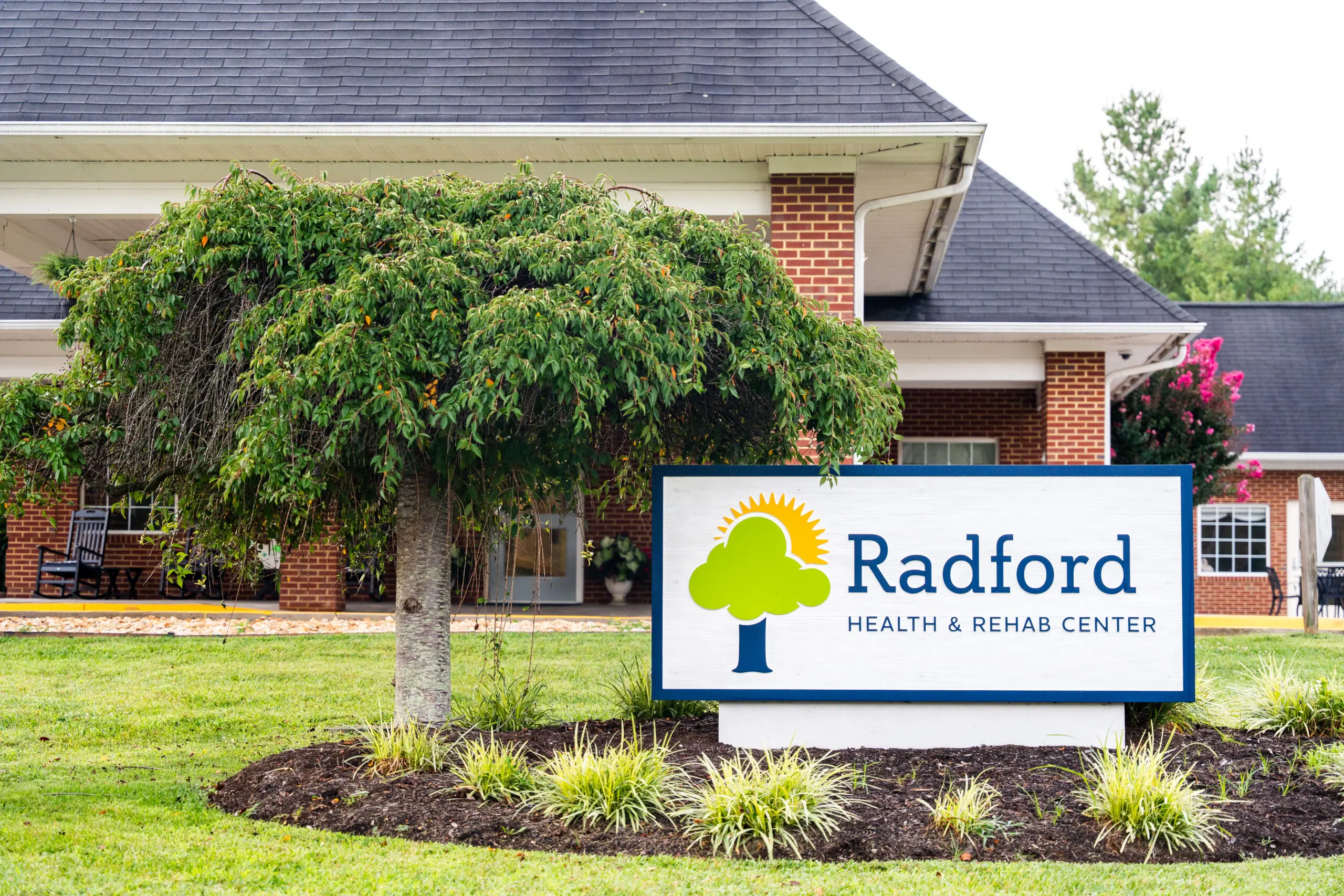 Photo of the Radford Health & Rehab logo/sign in a bed of mulch beside a tree outside the brick facility building