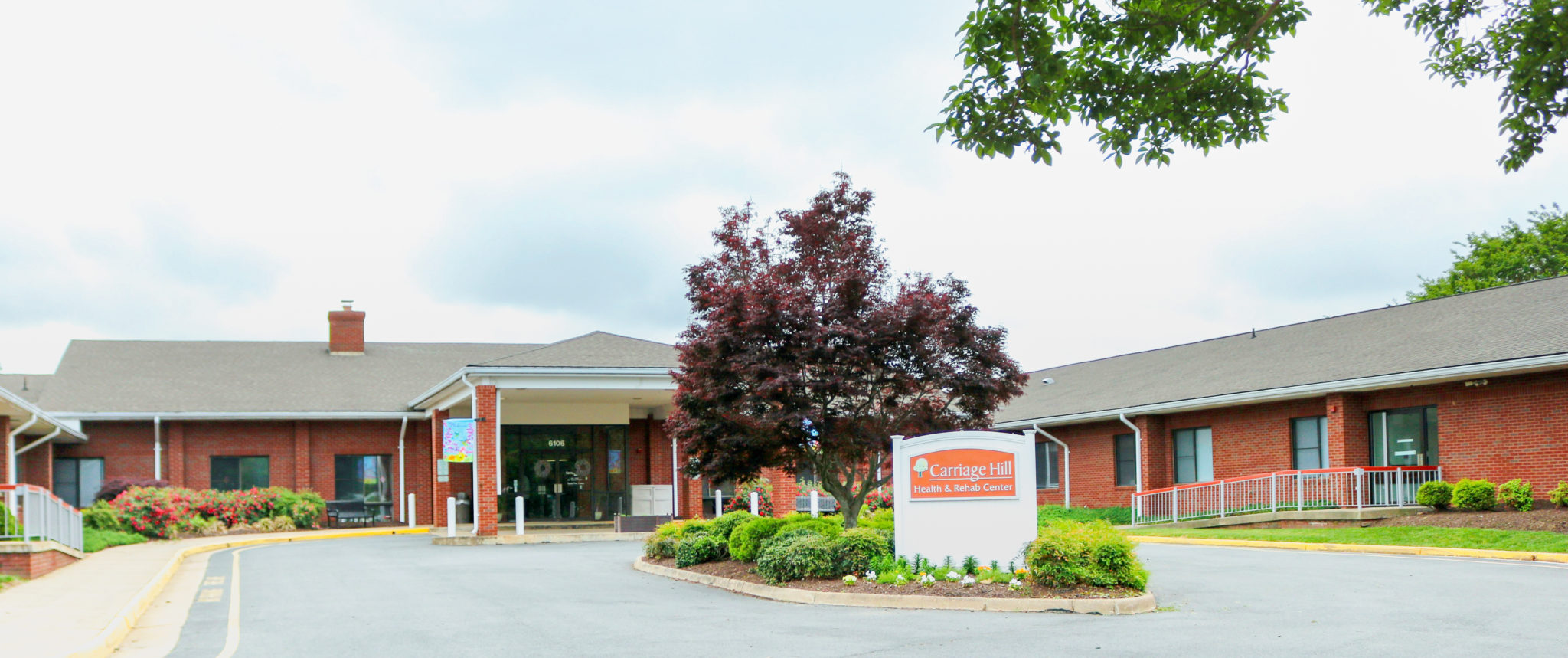 Exterior shot of a brick building, driving area, and a white brick sign that reads "Carriage Hill Health and Rehab Center"