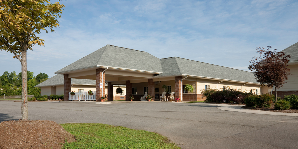 Exterior shot of a tan and brick building with a wide asphalt driveway and a covered porch area with rocking chairs