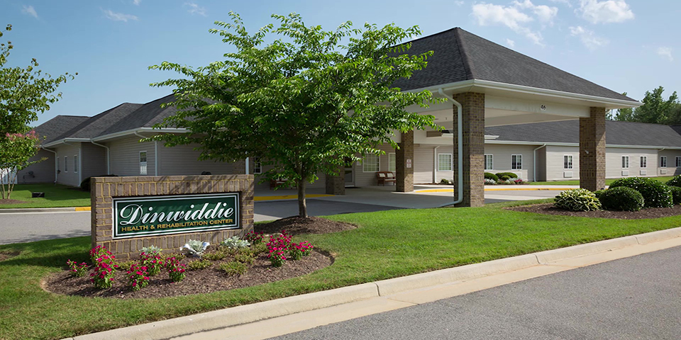 Exterior of a white and brick building and an asphalt driveway with a brick sign that reads "Dinwiddie Health & Rehab Center" in a bed of flowers in the foreground