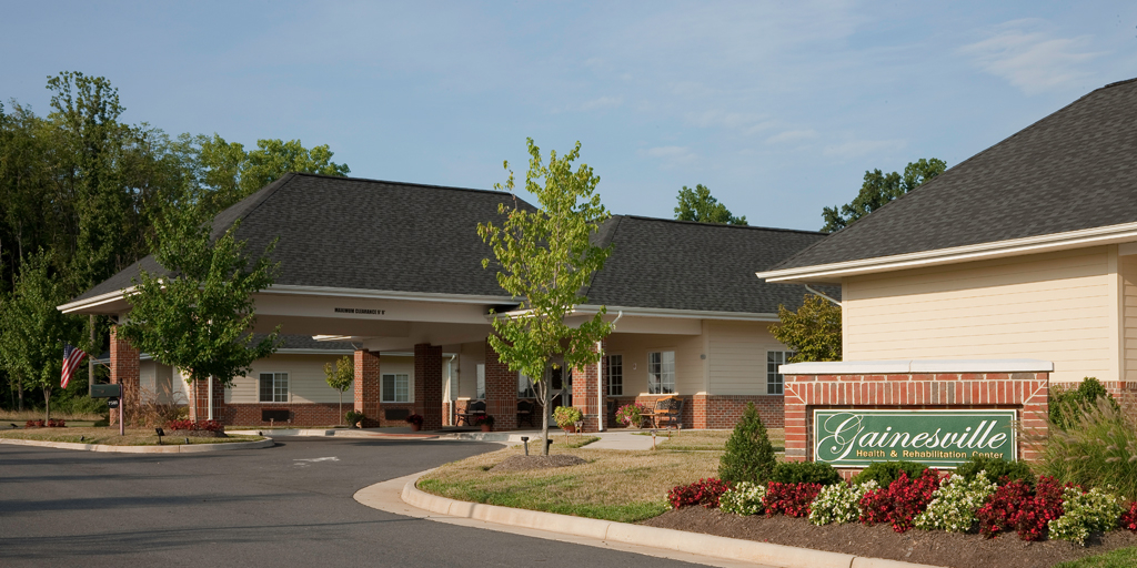 Exterior shot of brick and tan building and asphalt driveway with a brick sign that reads "Gainesville Health & Rehab Center" in a bed of flowers
