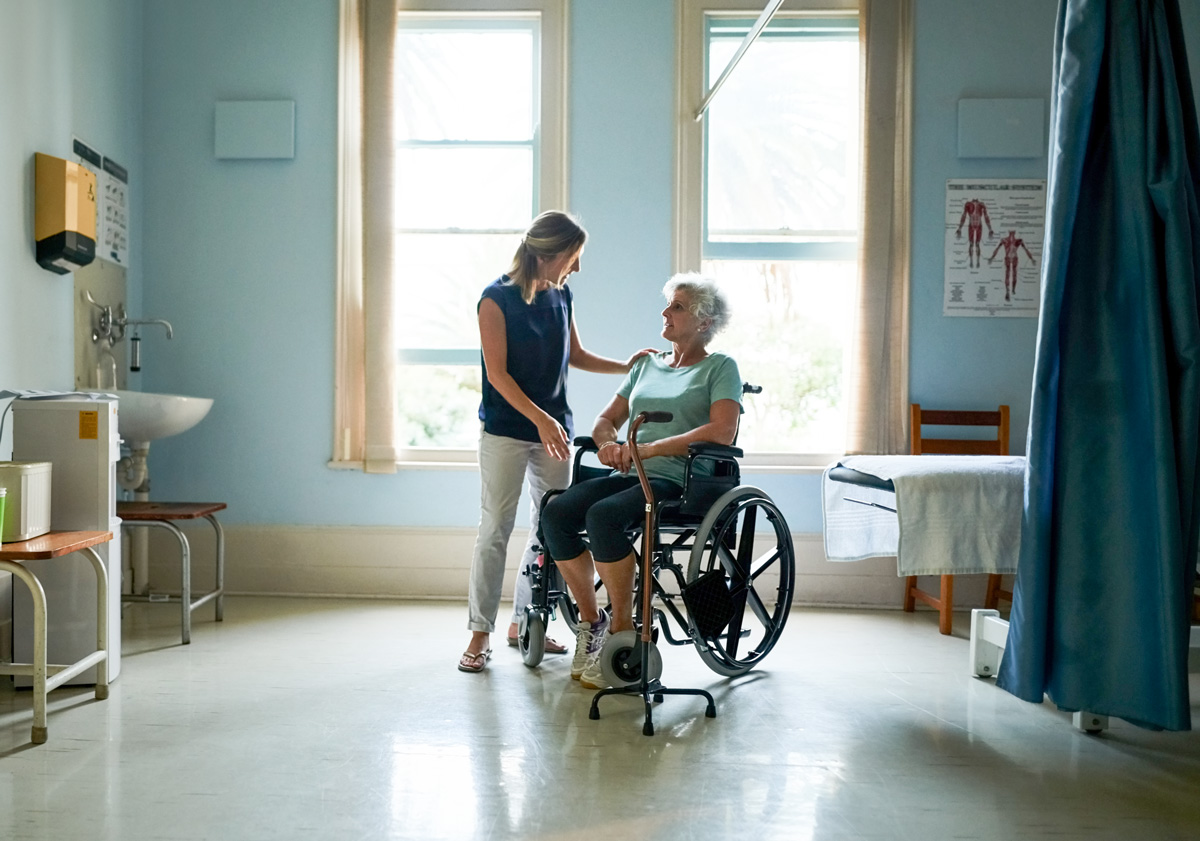 Blonde woman in blue top talks with older woman in a wheelchair while standing in a hospital room