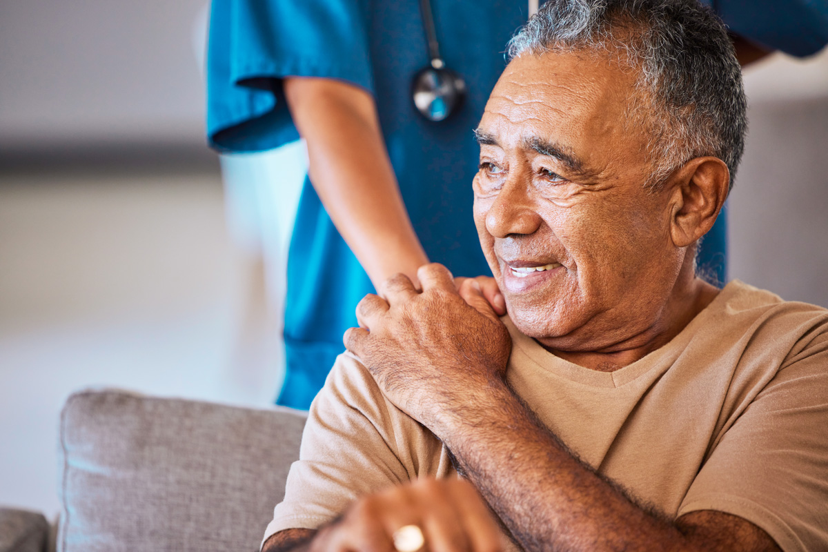 Older man in brown tshirt holds the hand of a nurse in blue scrubs who stands behind him with her hand on his shoulder
