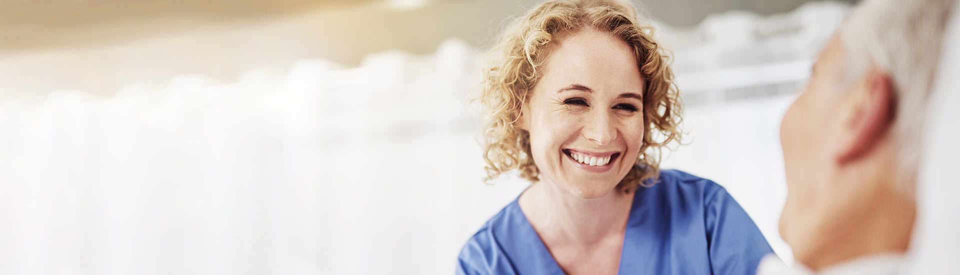 Curly-haired blonde nurse in blue scrubs smiles down at an elderly patient obscured in the foreground