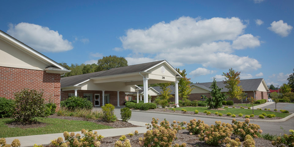 Exterior shot of a brick and white stone building, parking area, and garden beneath a blue sky