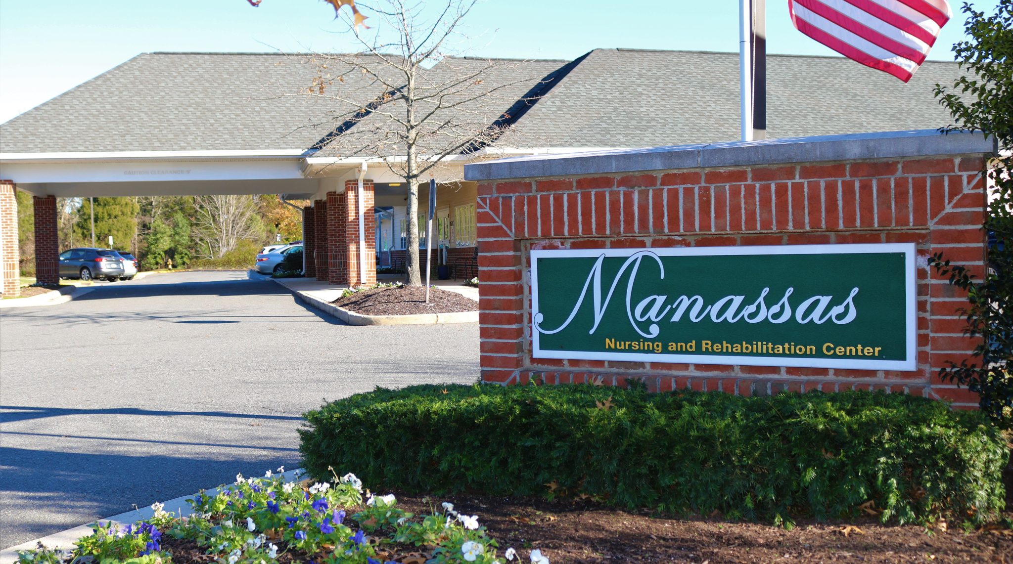 Close up shot of a brick sign beneath an American flag with "Manassas Nursing and Rehabilitation Center" on a green background