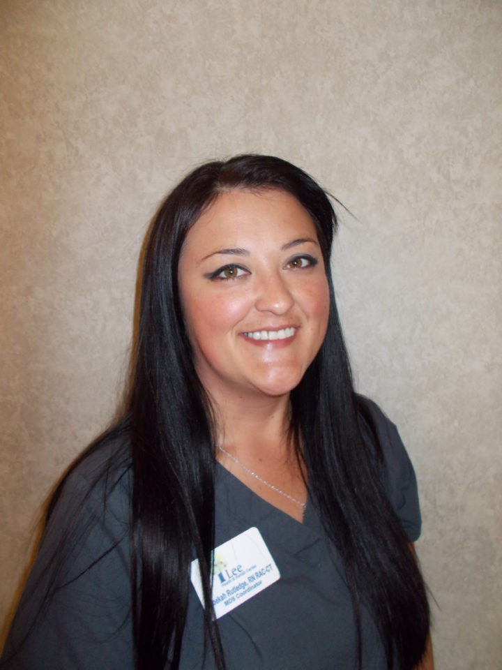 Woman with long dark hair and dark gray scrubs smiles for the camera in front of a beige background