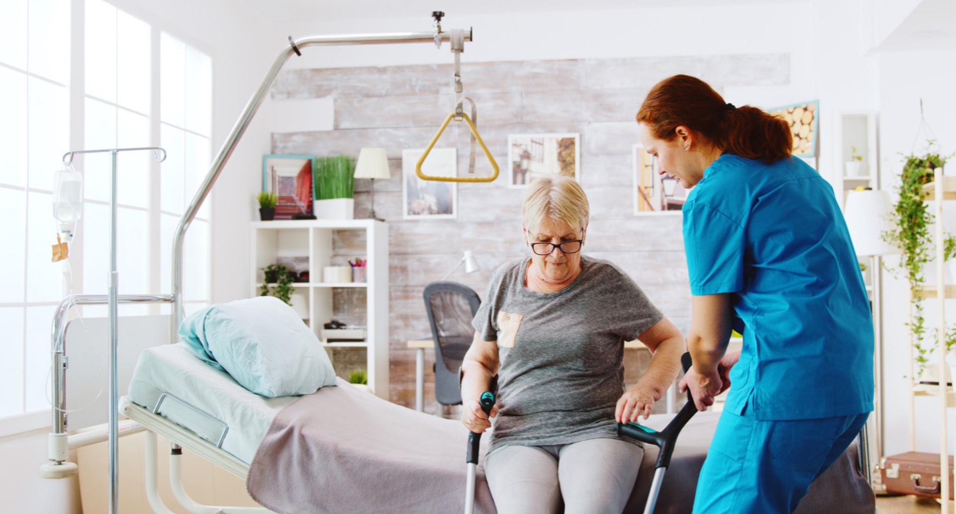 Red-haired female nurse in blue scrubs helps an older woman stand up from an examination table in a fancy exam room setting