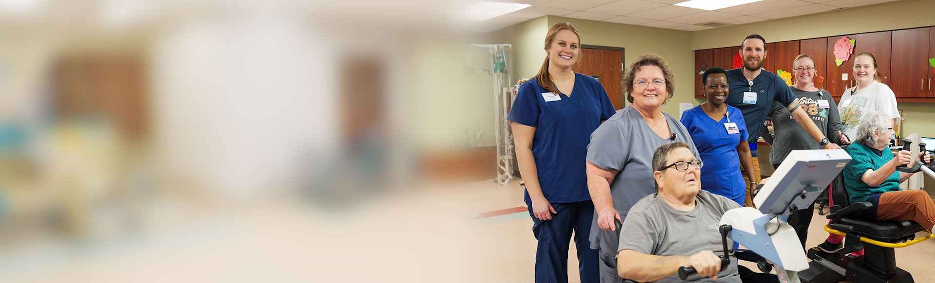 Group of male and female nurses stand behind patients on exercise machines in a medical room. The left half of the image is completely blurred