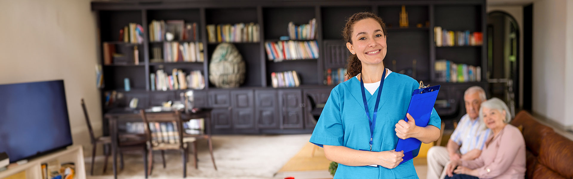 Brunette female nurse in blue scrubs and clipboard smiling in study with an elderly couple slightly blurred on the couch behind her