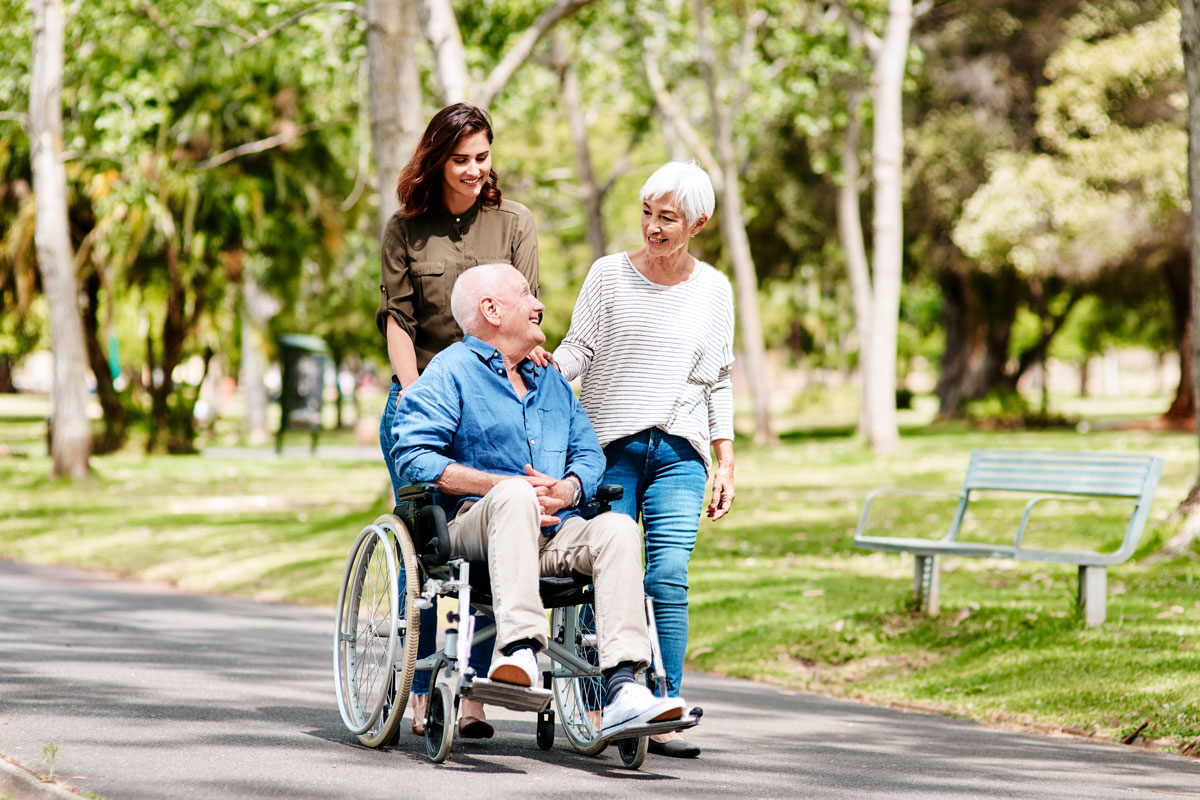 Brunette woman and white-haired woman push the wheelchair of an elderly man in a blue shirt through the park