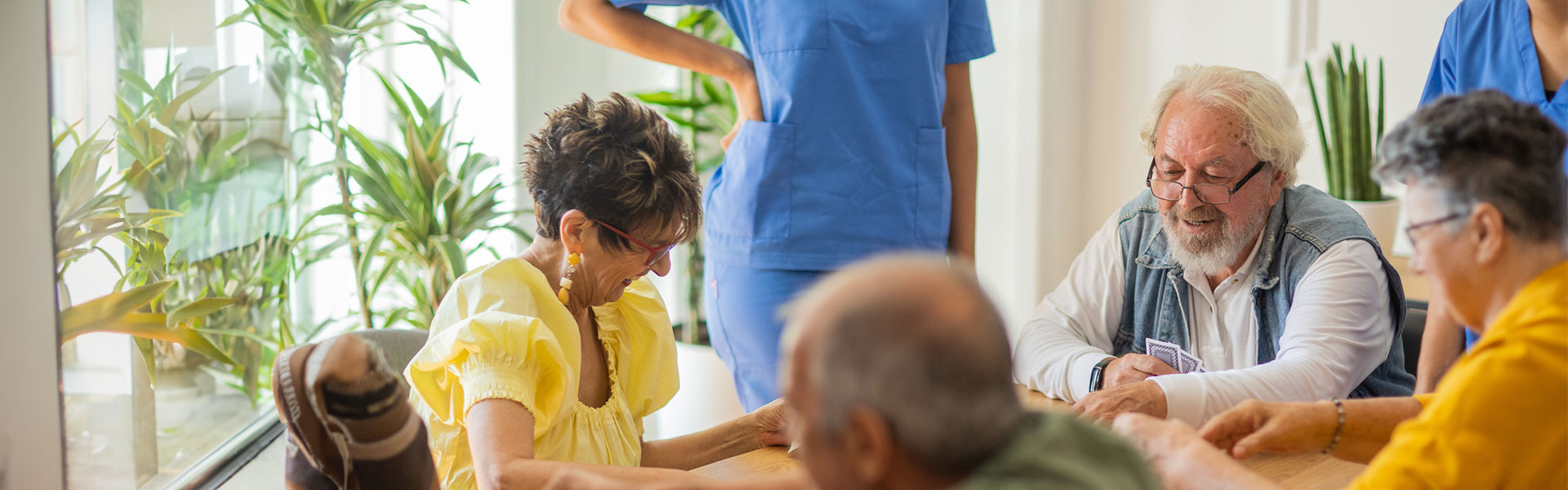 Group of older men and women sitting at a table, playing cards, and smiling while nurses in blue scrubs watch