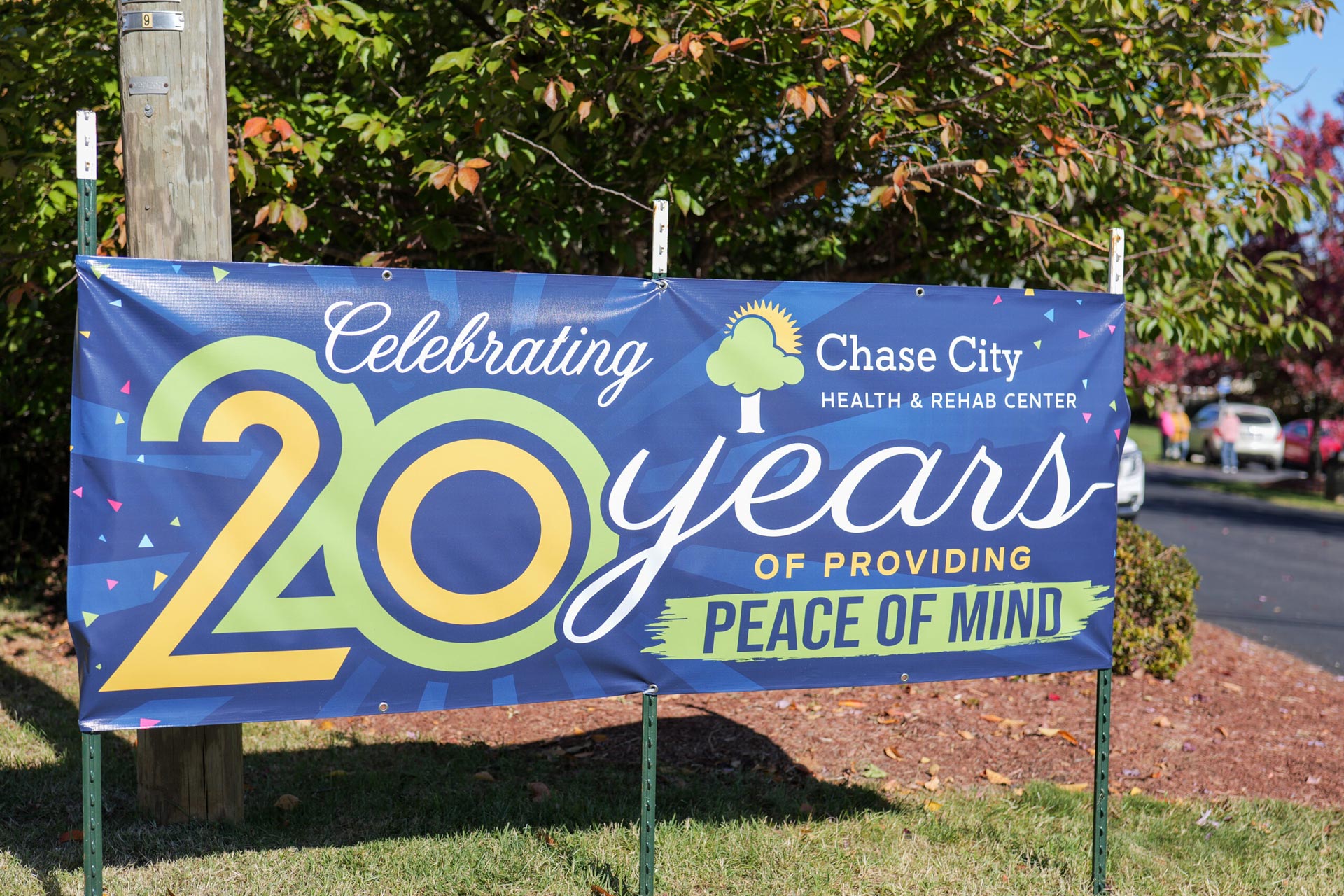 Green, yellow, and white text on a blue sunburst patterned banner alongside the road that reads "Celebrating 20 Years of Providing Peace of Mind" beneath the Chase City Health & Rehab Center logo