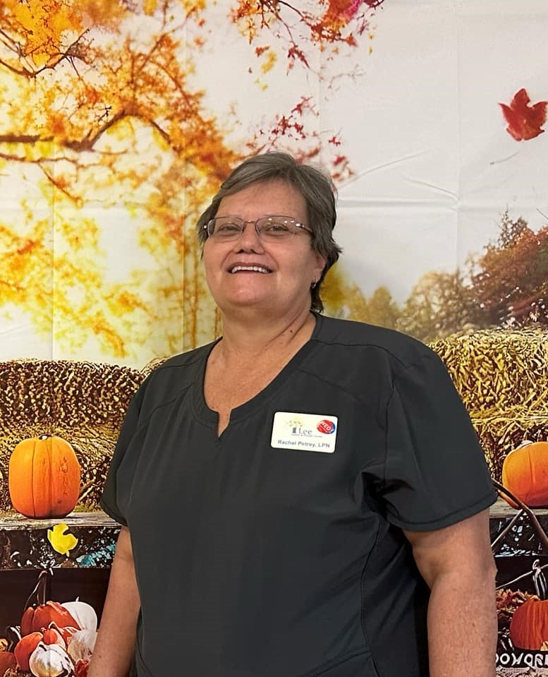 Older woman with gray/black hair and glasses wears dark gray scrubs and smiles for the camera while standing in front of a fall background