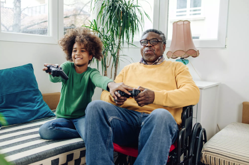 Young boy and an older man in a wheelchair hold video game controllers and play together in a sunlit room