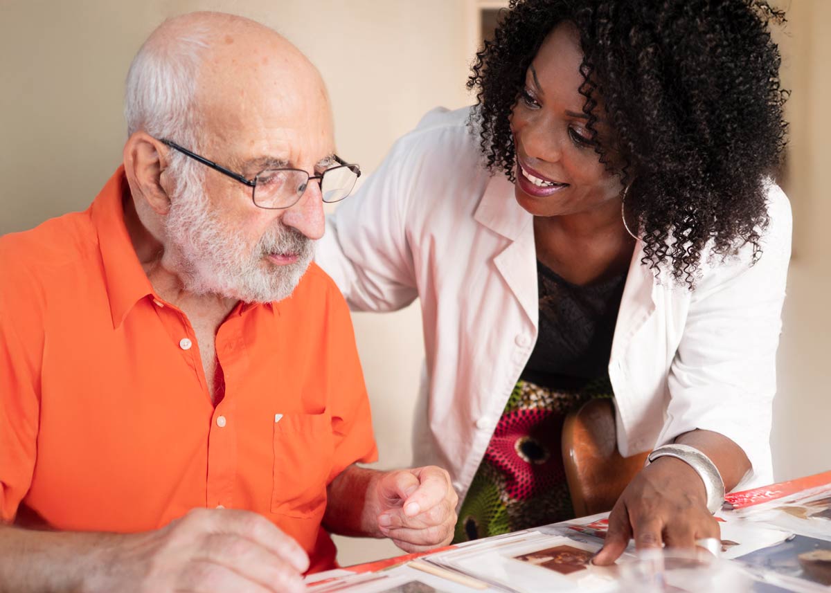Woman in a white lab coat stands beside old man in an orange shirt looking at papers on a table