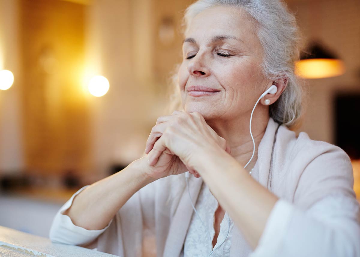 Older woman in a white blouse and cream colored shawl wears white wired headphones and clasps hands under chin with eyes closed in a room lit with warm yellow light