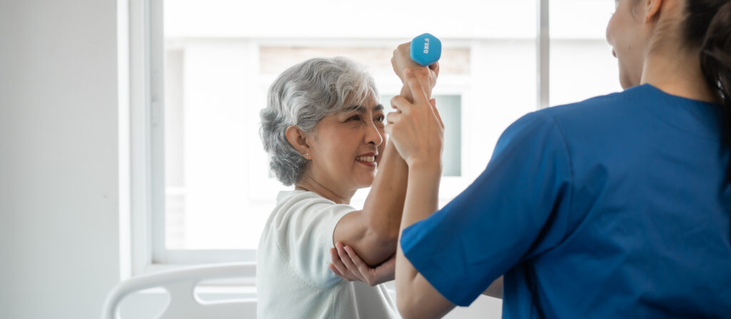 Young physiotherapist helping senior mature asian woman grey hair work out with dumbbells, to recover from injury at health centre in physical therapy session.