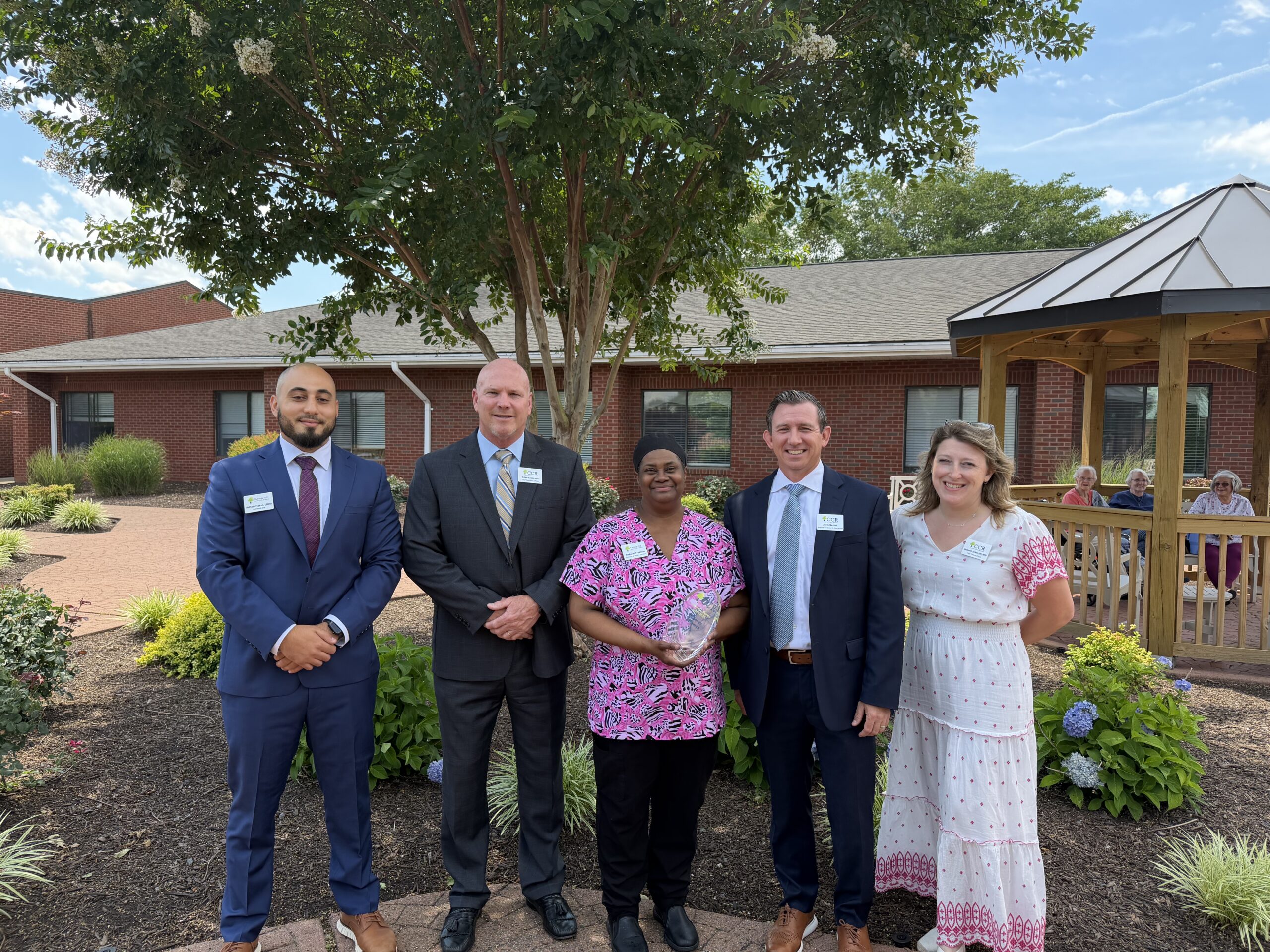 Three men in suits, a female nurse in pink scrubs, and a woman in a dress smile together in a garden outside a brick building