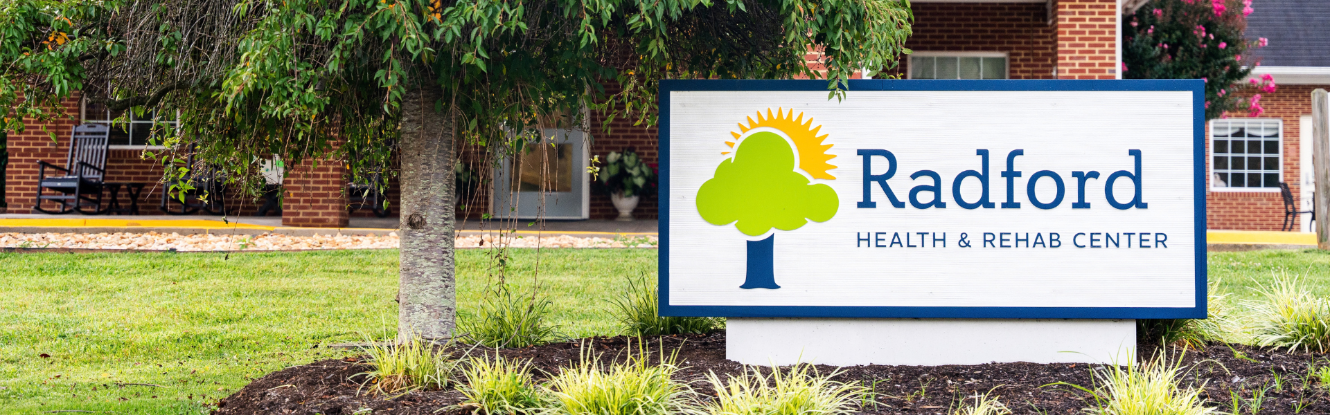 Photo of the Radford Health & Rehab logo/sign in a bed of mulch beside a tree outside the brick facility building
